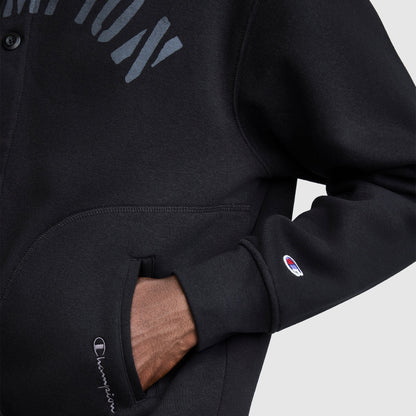 A person wears the Champion Fleece Baseball Jacket in black, with one hand tucked into the pocket. The signature Champion logo appears on both the sleeve and pocket, highlighting its vintage athletic style inspired by classic varsity jackets.