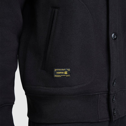 Close-up of the lower front half of the Champion Fleece Baseball Jacket in black, featuring side pockets, snap buttons, and a small yellow and black Champion logo tag near the waist. This vintage athletic-inspired piece highlights classic varsity jacket details.