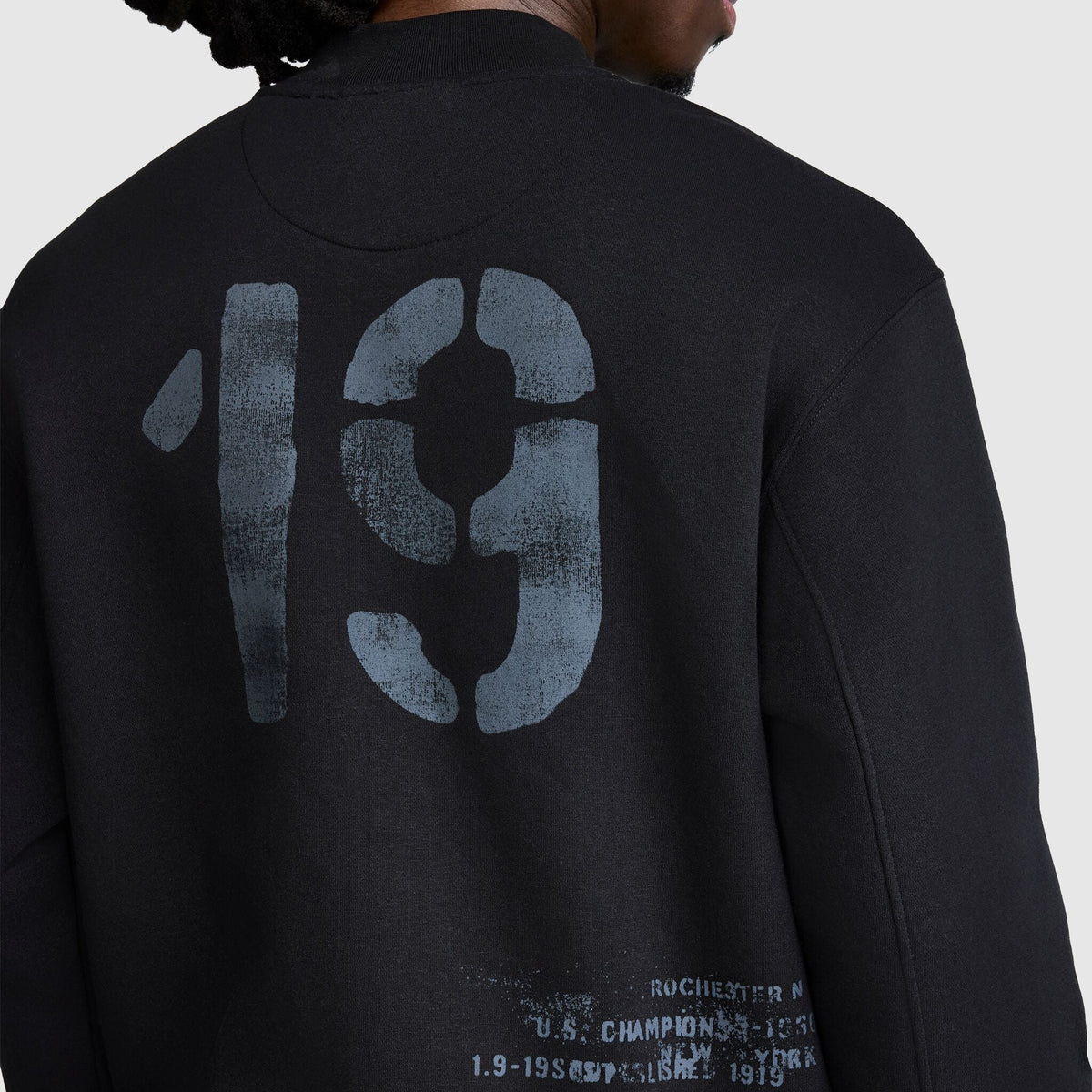 A person wearing the Champion Fleece Baseball Jacket in black, featuring a large, weathered number 19 on the back and faded text near the bottom that partially reads “U.S. CHAMPIONSHIPS” and “ROCHESTER N.Y.,” giving it a vintage athletic feel.