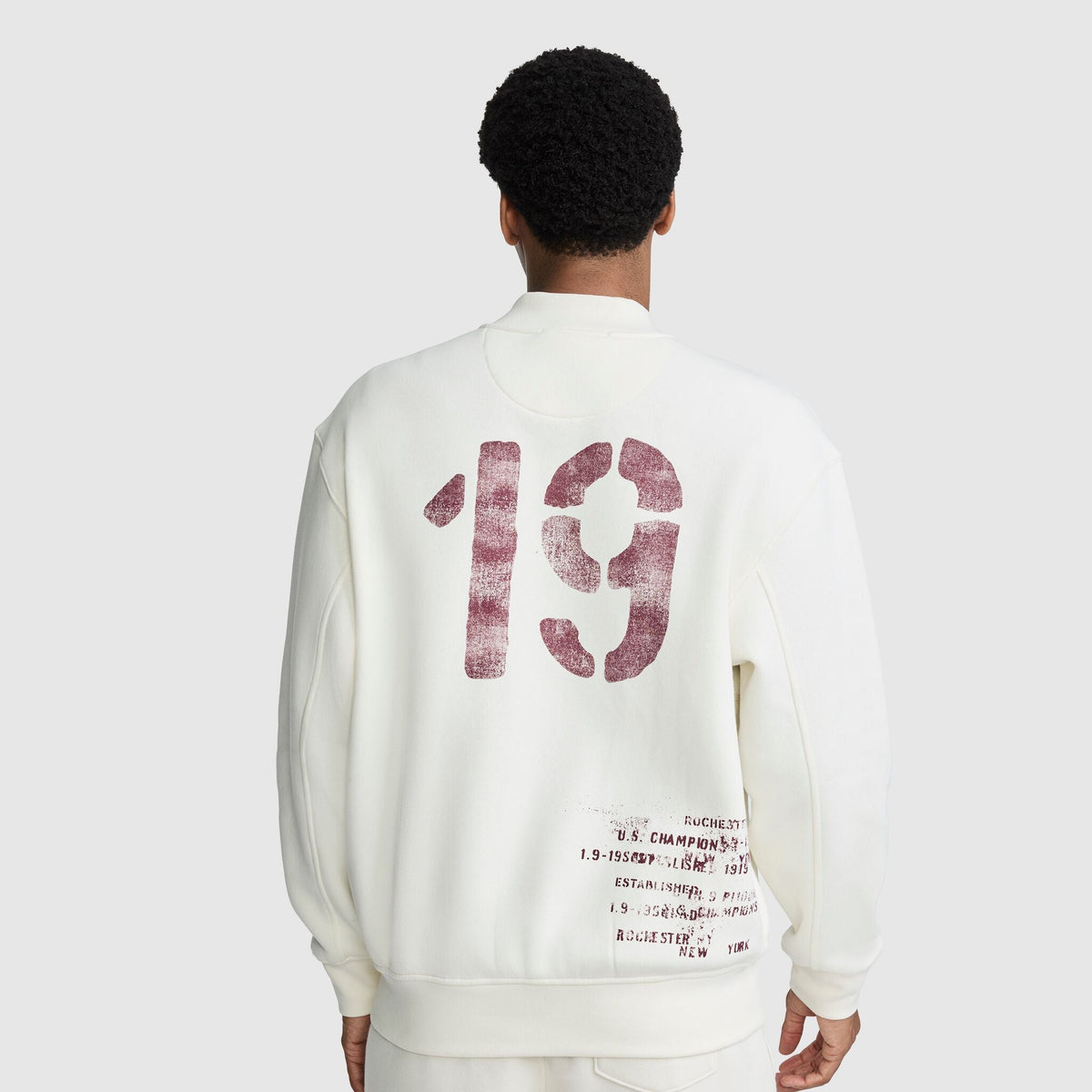 Back view of a person wearing a cream-colored Champion Fleece Baseball Jacket featuring large red 19 branding on the back and additional printed text near the hem.
