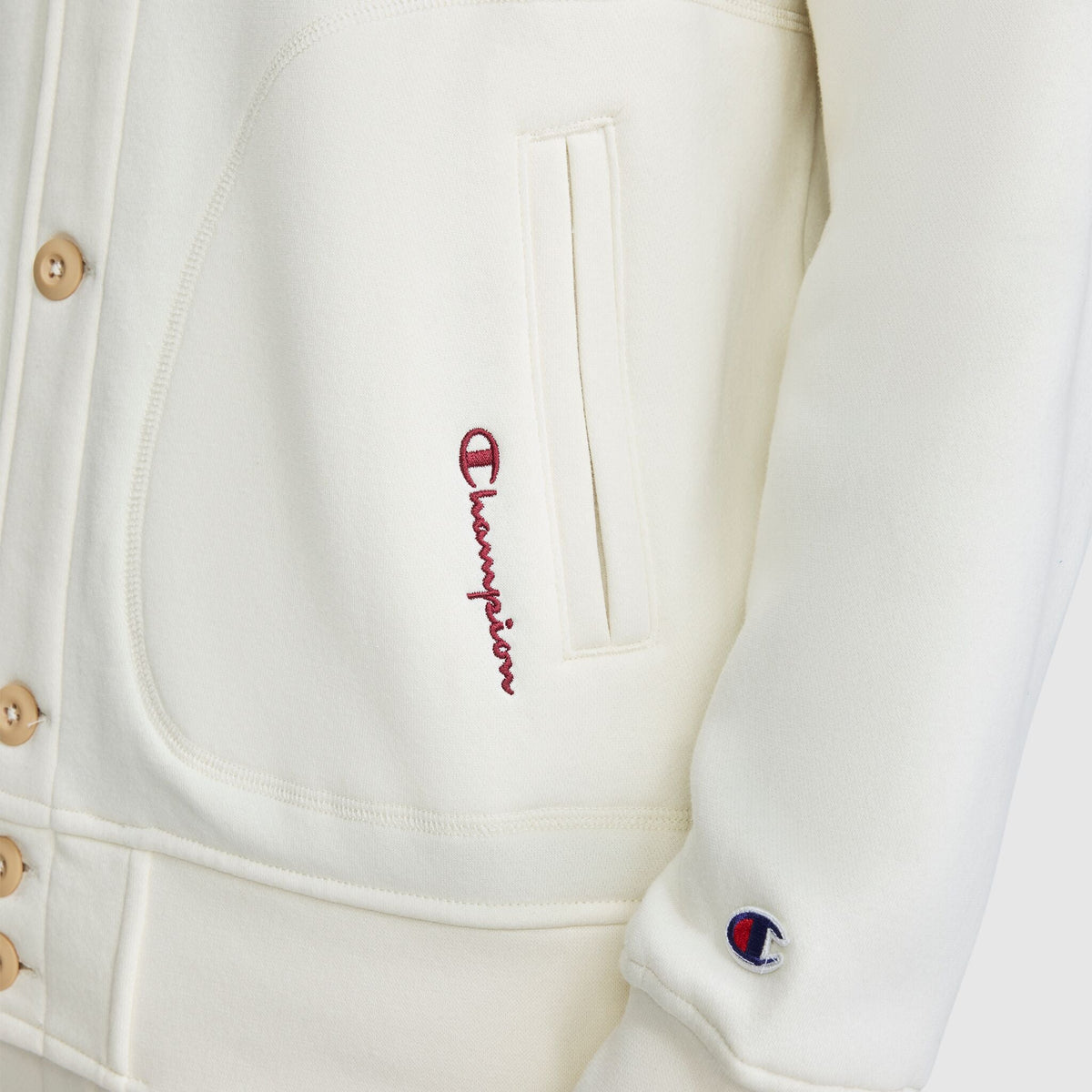 Close-up of a cream-colored Champion Fleece Baseball Jacket with a small embroidered logo on the sleeve and red cursive text near the pocket.