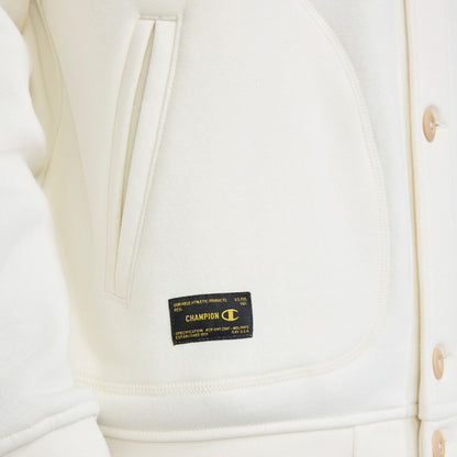 Close-up of a cream-colored Champion Fleece Baseball Jacket with a button front and a black rectangular tag featuring gold text and a logo on the lower pocket area.