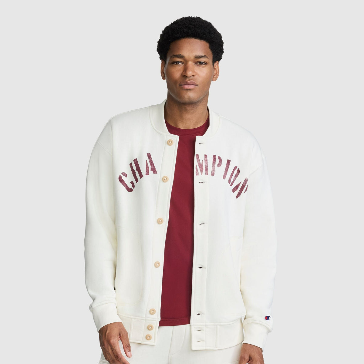 A man wears an unbuttoned, cream-colored Champion Fleece Baseball Jacket with maroon lettering across the chest over a matching maroon t-shirt.