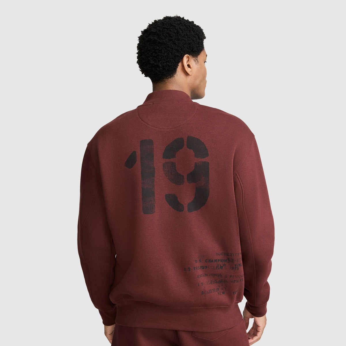 A person viewed from behind wearing a maroon Champion Fleece Baseball Jacket, featuring a large black 19 graphic and smaller printed text on the back.