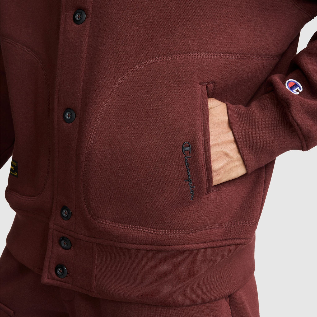 Close-up of a maroon Champion Fleece Baseball Jacket with a hand in the front pocket and a logo embroidered on the sleeve.