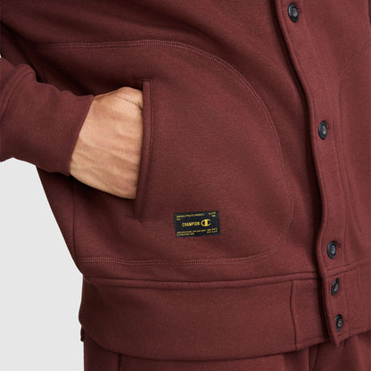 Close-up of a hand in the pocket of a maroon Champion Fleece Baseball Jacket with a rectangular brand patch near the hem.