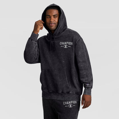 A man wearing a black Mineral Wash Pullover Hoodie, C Patch Logo with the word CHAMPION and a logo printed on the chest.