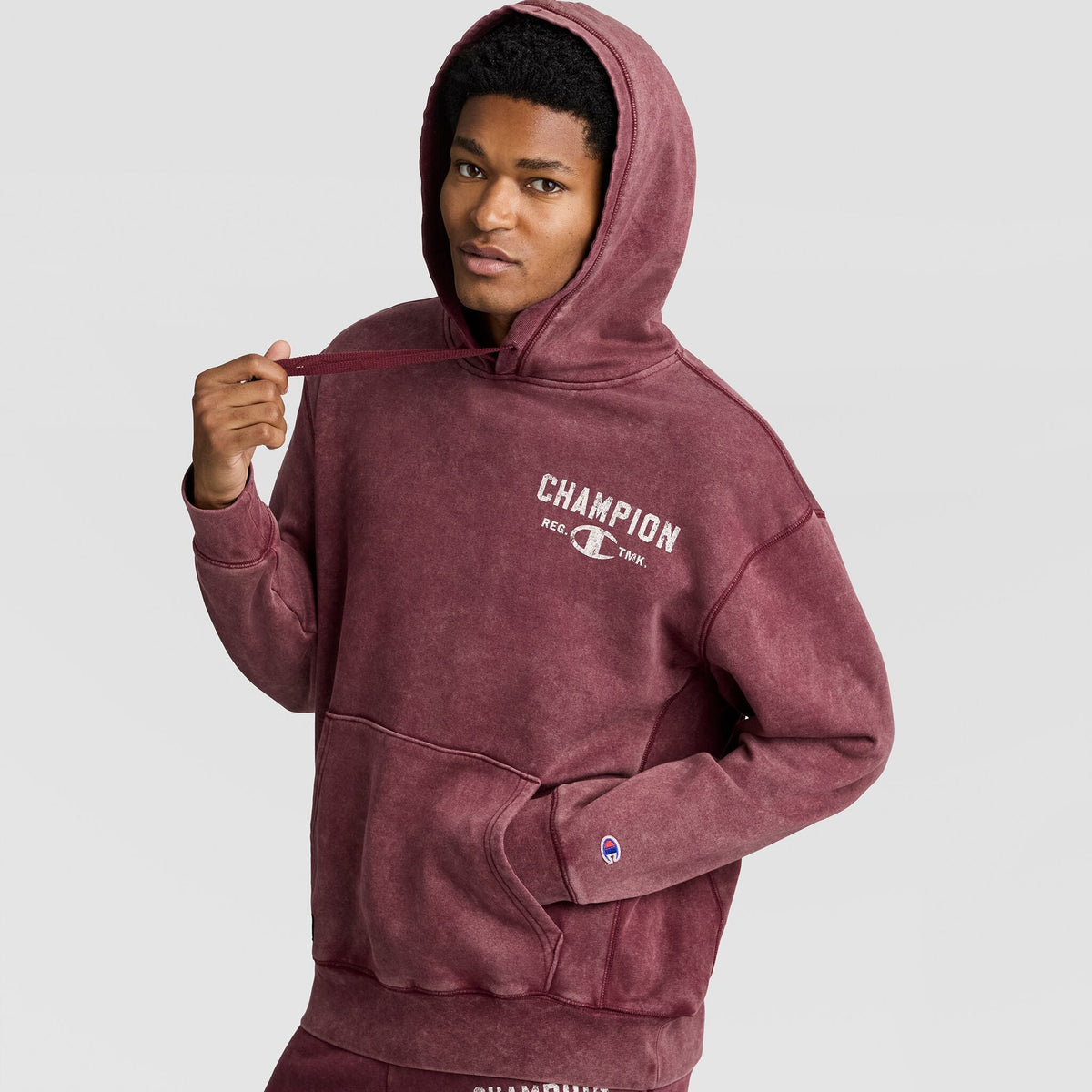 A man wearing a burgundy Mineral Wash Pullover Hoodie, C Patch Logo with a white logo on the chest and a small patch on the sleeve.
