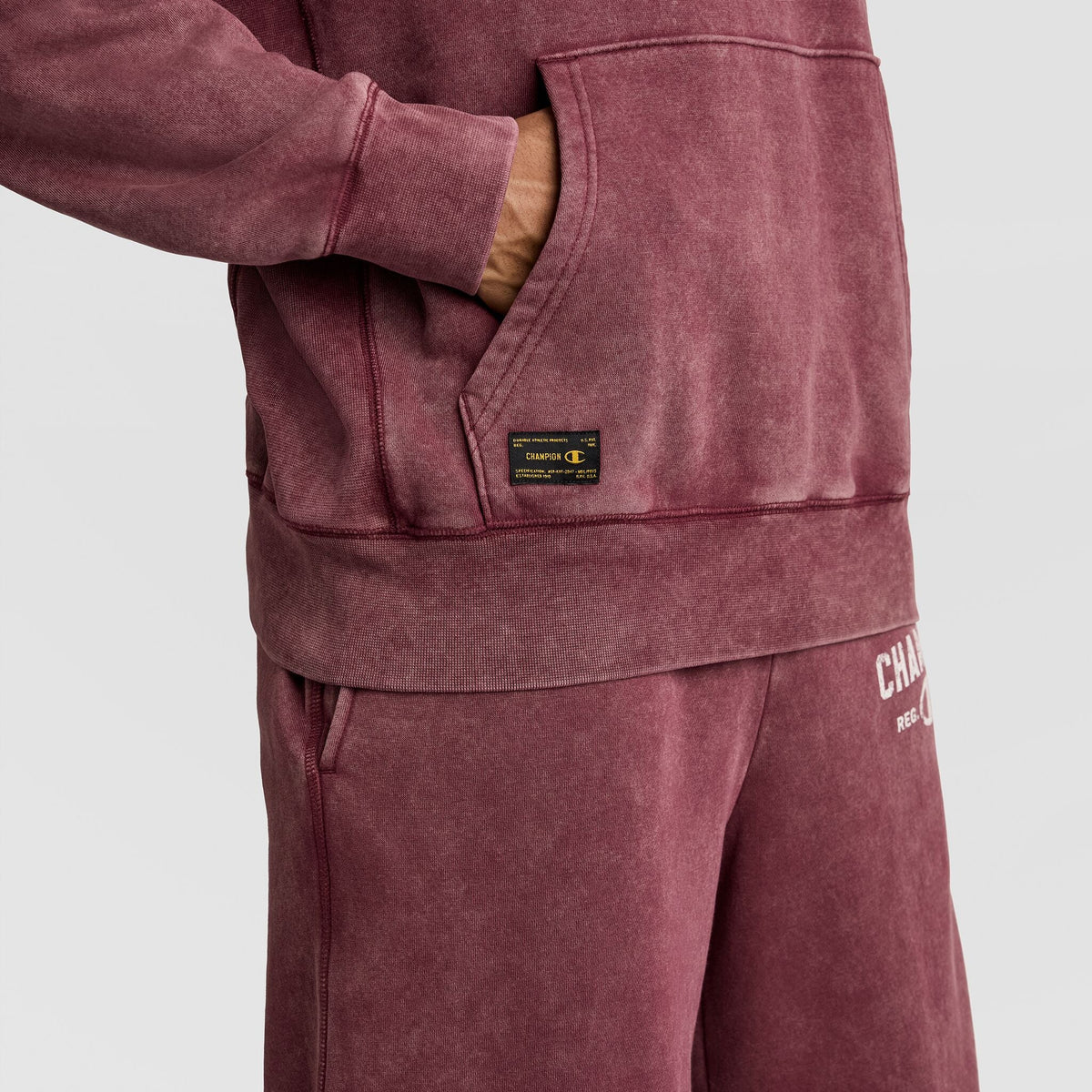 A person wearing a dark red Mineral Wash Pullover Hoodie, C Patch Logo with a rectangular patch on the front kangaroo pocket and matching sweatpants.