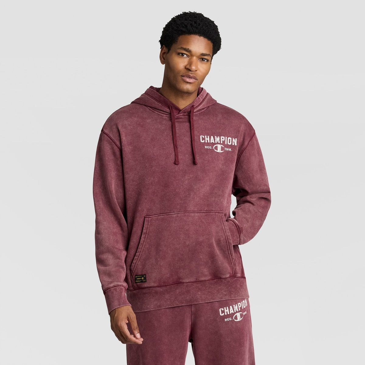 A man wearing a burgundy Mineral Wash Pullover Hoodie, C Patch Logo and matching sweatpants, both featuring printed logos.