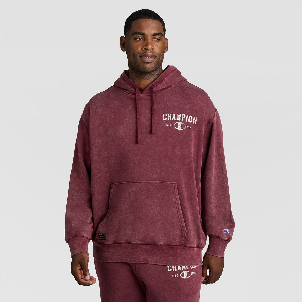 A man wearing a burgundy Mineral Wash Pullover Hoodie, C Patch Logo, featuring a graphic logo on the chest and a small patch on the sleeve.