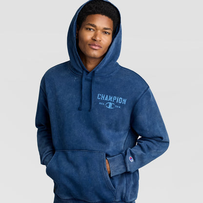 A person wearing a blue mineral wash Champion Mineral Wash Pullover Hoodie, C Patch Logo featuring a kangaroo pocket and a distressed graphic on the chest.
