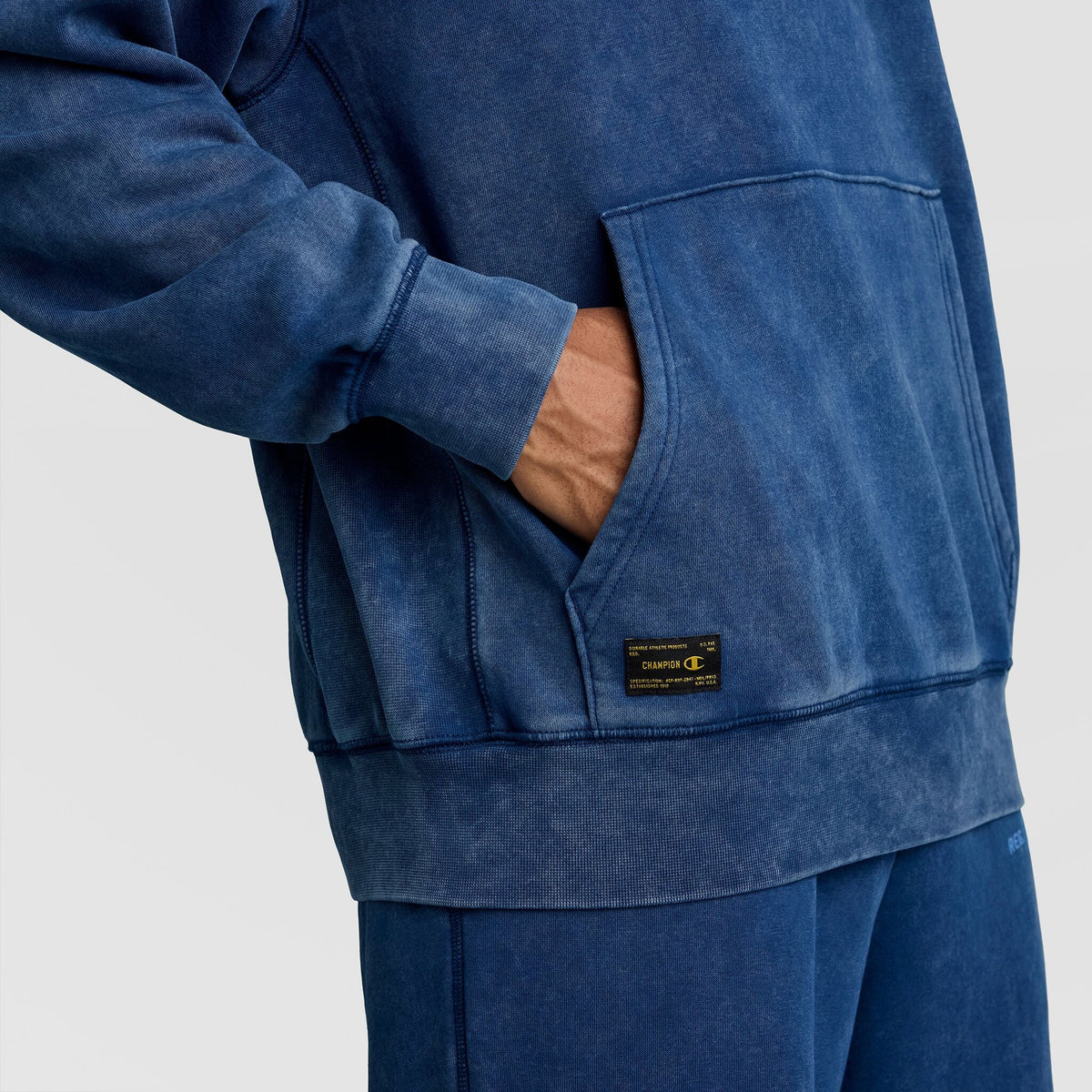 Close-up of a blue Mineral Wash Pullover Hoodie, C Patch Logo by Champion, showing a hand in the front pouch pocket and the rectangular logo patch near the hem.