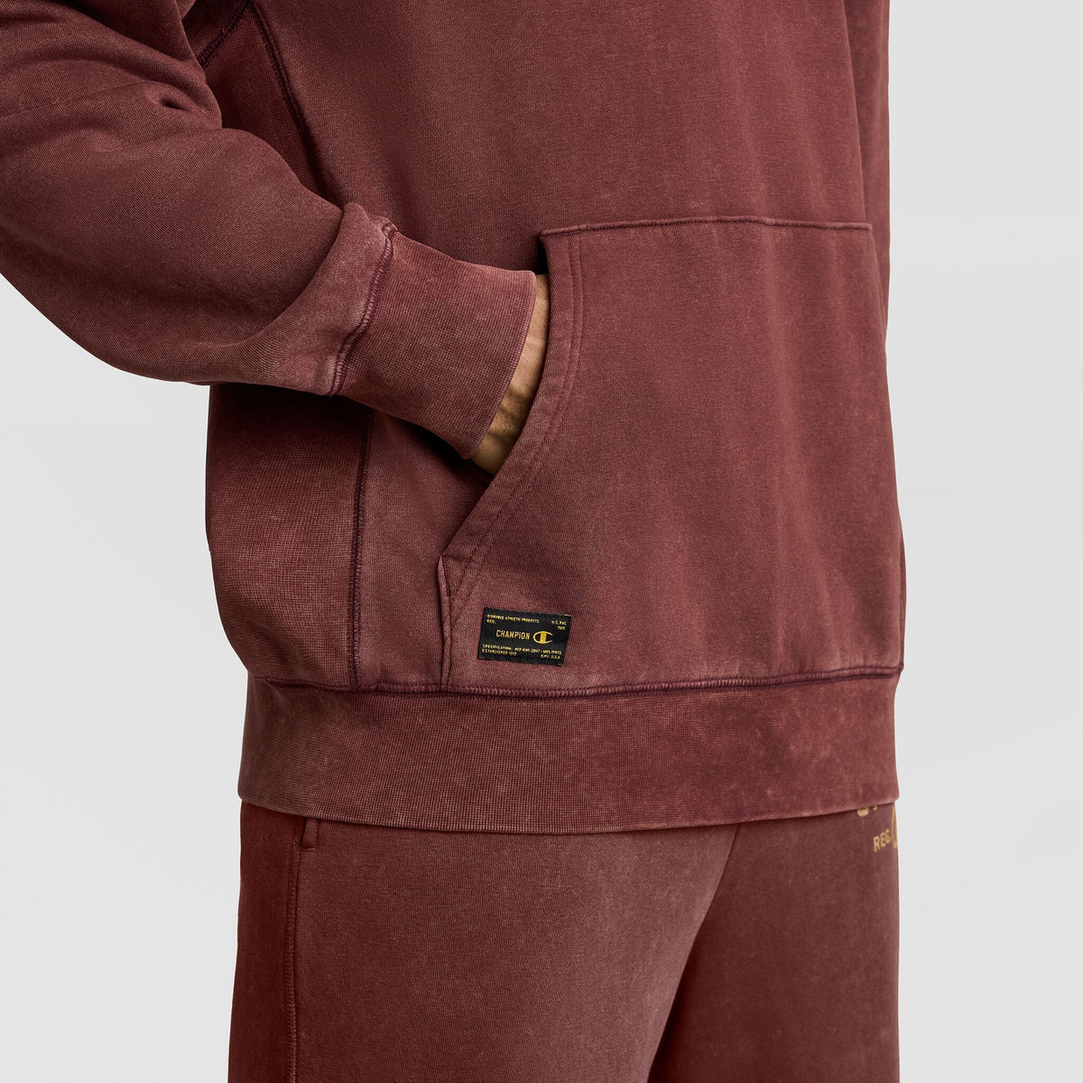 A person wearing a vintage dark red Champion Mineral Wash Pullover Hoodie, C Patch Logo, with a hand in the front pouch pocket, which features a rectangular woven patch label.