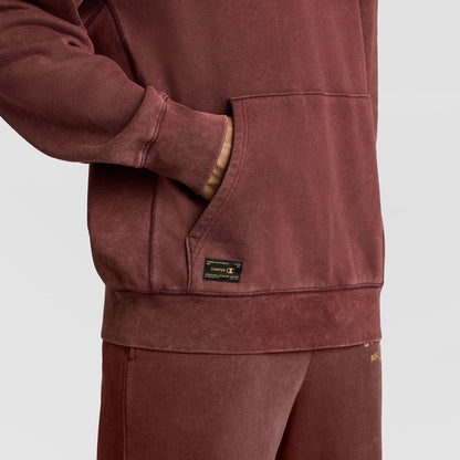 A person wearing a vintage dark red Champion Mineral Wash Pullover Hoodie, C Patch Logo, with a hand in the front pouch pocket, which features a rectangular woven patch label.