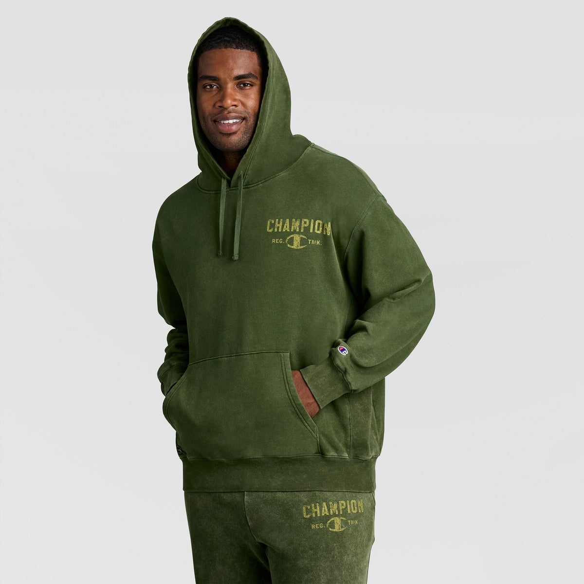 A man wearing a heavy olive green Mineral Wash Pullover Hoodie, C Patch Logo featuring a distressed chest logo and a small sleeve patch.