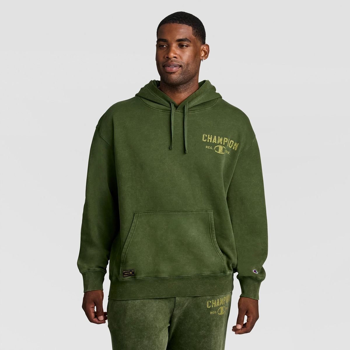 Man wearing an olive green, heavy mineral wash Champion Mineral Wash Pullover Hoodie, C Patch Logo featuring a distressed-look logo on the chest and a small patch on the pocket.