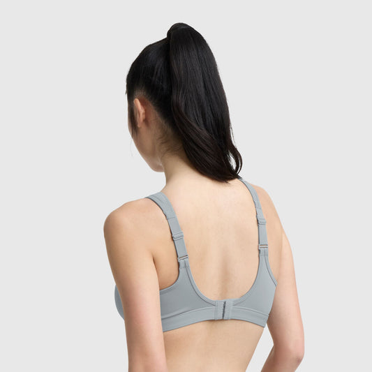 A woman with long dark hair in a high ponytail is seen from the back, wearing the Champion Spot Comfort High-Impact Sports Bra (C Logo) in light gray with adjustable straps, against a plain light background.