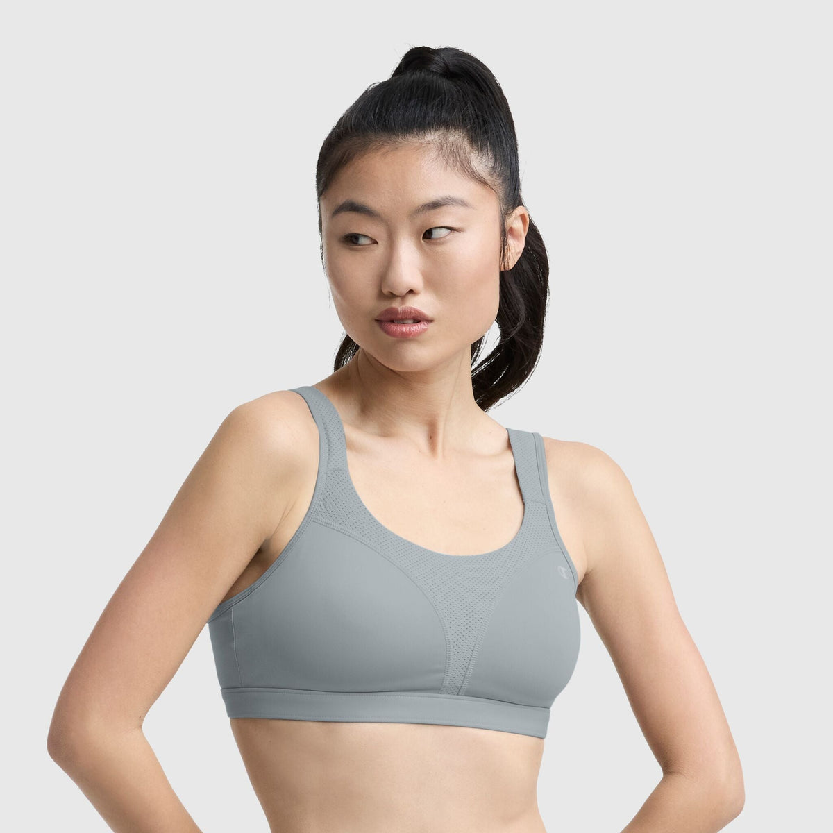 A woman with long dark hair in a high ponytail wears the Champion Spot Comfort High-Impact Sports Bra, C Logo in light gray, looking to her left against a plain light gray background.