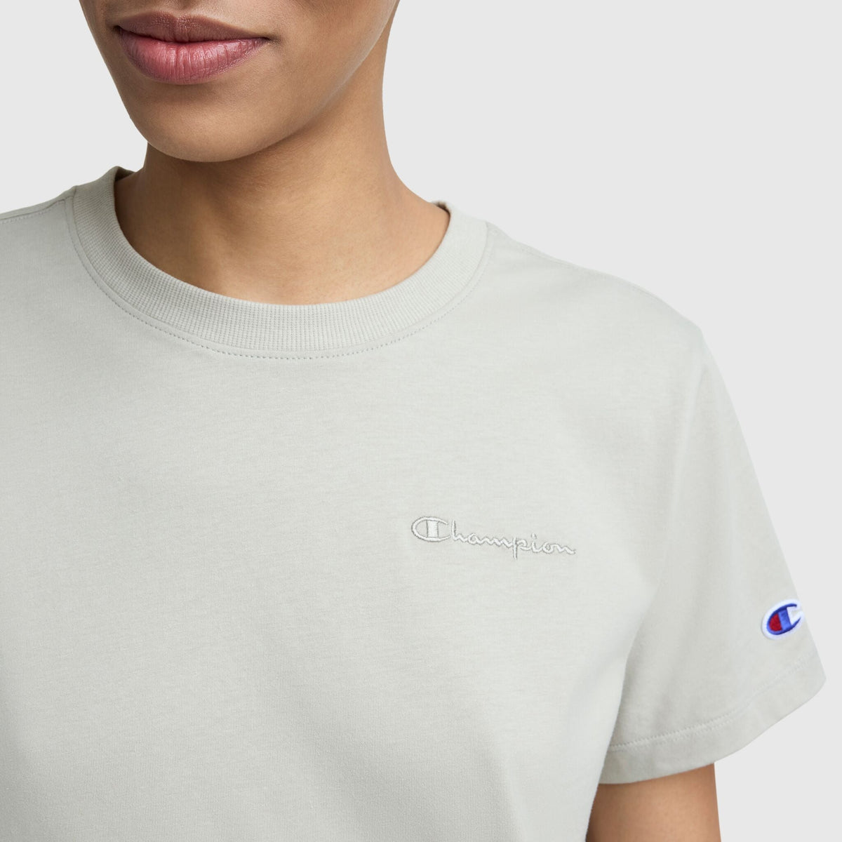 A person wears a light gray women's Champion Classic T-Shirt with tonal left chest script and a sleeve logo patch, shown from the shoulders up against a plain background.