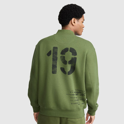 A person is shown from the back wearing Champion’s Fleece Baseball Jacket in olive green, paired with matching pants. Channeling a vintage athletic vibe, the jacket features a bold black “19” and smaller text printed near the lower back, echoing classic varsity jacket design.