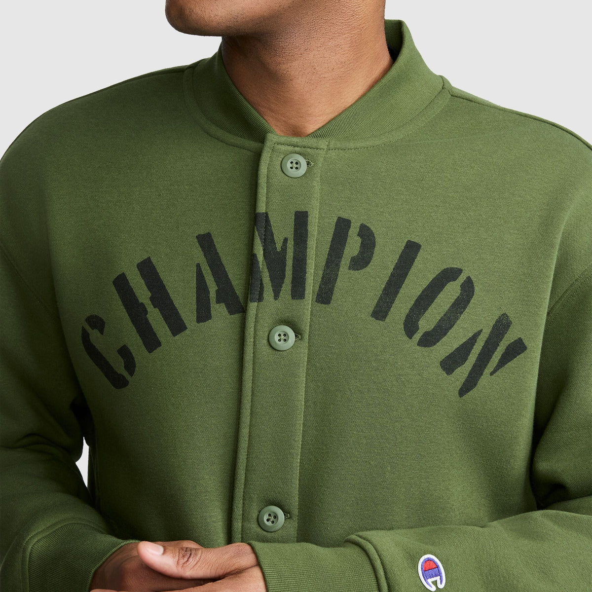 A person wearing a green Champion Fleece Baseball Jacket featuring CHAMPION in bold black letters across the chest and a small Champion logo on the sleeve, giving off a vintage athletic vibe.
