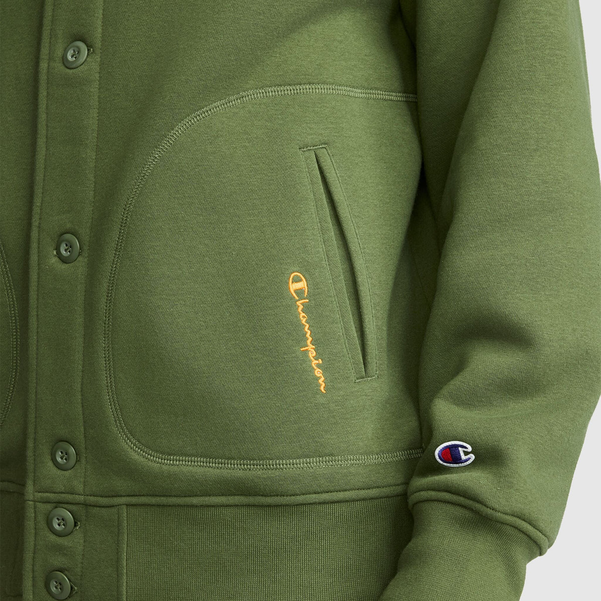 Close-up of a green vintage-inspired Champion Fleece Baseball Jacket featuring a front pocket with a yellow Champion logo, plus a small Champion C patch on the sleeve. The jacket is designed with visible buttons and detailed stitching, echoing the classic varsity jacket style.