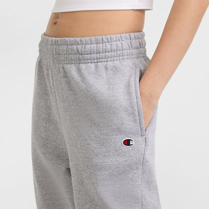 A person wears Champion Powerblend Oversized Sweatpants featuring a C logo on the left hip, elastic waistband, light gray color, one hand in the pocket, and part of a white crop top showing.