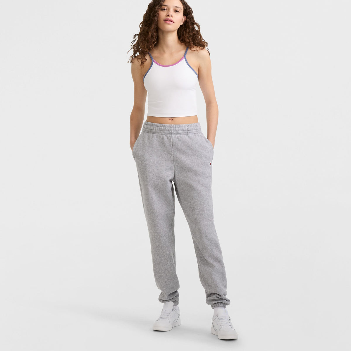 A person with curly hair, wearing a white crop tank top with purple trim, Champion Powerblend Oversized Sweatpants (C Logo), and white sneakers stands with hands in pockets against a light background.