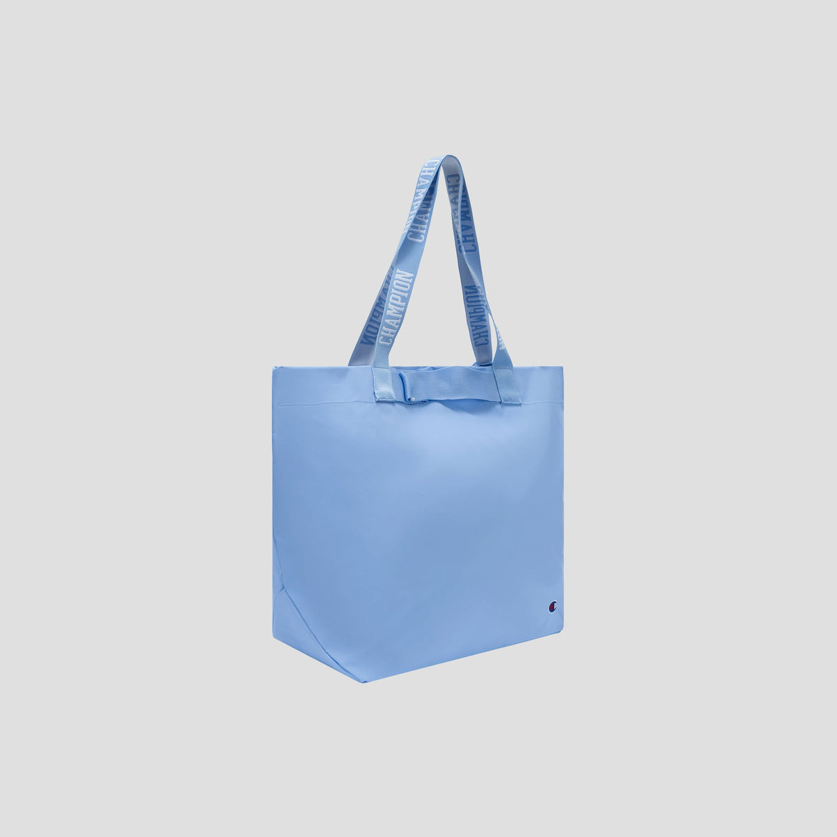 The Champion Miller Coated Shopper Tote is a light blue, weather-resistant bag featuring long, flat handles and bold white text. Its spacious and minimalist design is accented by a small red and black detail near the bottom corner. The bag is pictured against a plain light gray background.