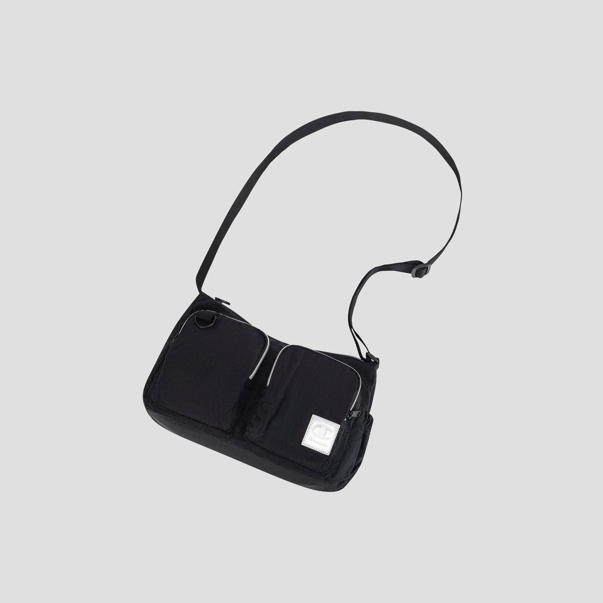 The Champion Quartz Small Messenger Bag is a lightweight nylon crossbody in black, featuring two front zip pockets, an adjustable strap, and a small white rectangular patch on the lower right front, displayed against a plain light gray background.