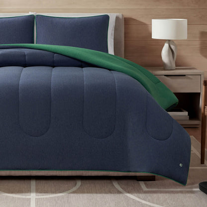 A bed featuring the Champion Premium Reversible Solid Jersey Comforter Set in navy blue and green, paired with matching pillows, alongside a wooden nightstand topped with a white lamp, and a light-colored rug adorned with a geometric pattern.