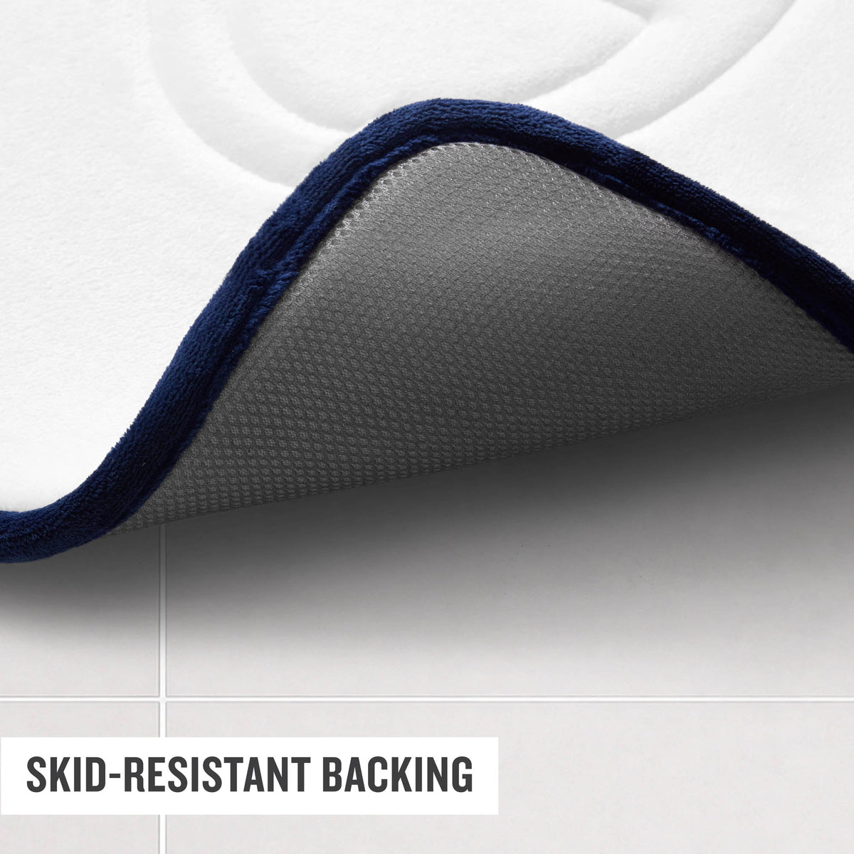 A close-up of the corner of a Champion Quilted Logo Memory Foam Bath Mat lifted up, revealing its textured, gray skid-resistant backing on a tile floor. The image also features text that reads SKID-RESISTANT BACKING.