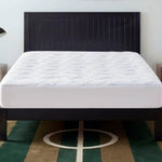 A neatly made bed with a white, antimicrobial and stain-resistant mattress is topped by the Champion Cooling Knit Mattress Pad and features a dark wooden headboard, all situated on a green and brown rug with a white geometric pattern.