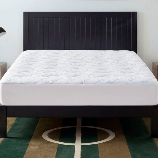A neatly made bed with a white, antimicrobial and stain-resistant mattress is topped by the Champion Cooling Knit Mattress Pad and features a dark wooden headboard, all situated on a green and brown rug with a white geometric pattern.