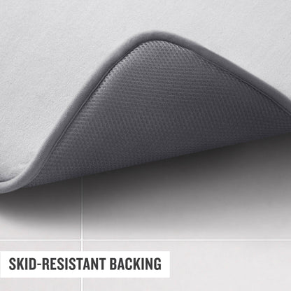 A Champion Logo Patch Memory Foam Bath Mat in gray is displayed with one corner lifted to reveal its textured, skid-resistant backing. The mat rests on light-colored tiles, and text on the image reads “SKID-RESISTANT BACKING.”.