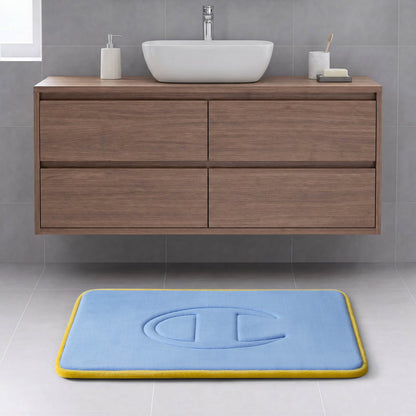 A modern bathroom with a floating wooden cabinet, white sink, soap dish, and a Champion Quilted Logo Memory Foam Bath Mat in blue with a yellow border and embossed design on a light gray tiled floor.