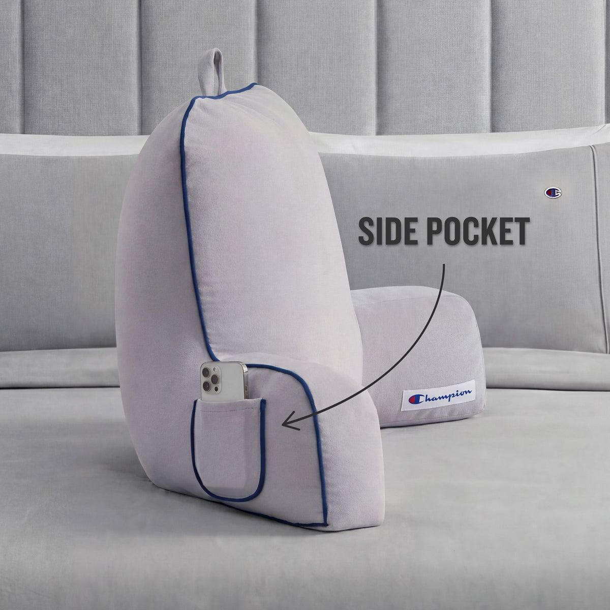 A light gray Champion Faux Wool Backrest Pillow with supportive arms and blue piping rests on a gray bed. Made from soft faux wool, the pillow includes a small side pocket—highlighted with an arrow and the label “SIDE POCKET”—holding a smartphone. The Champion logo patch is also visible.