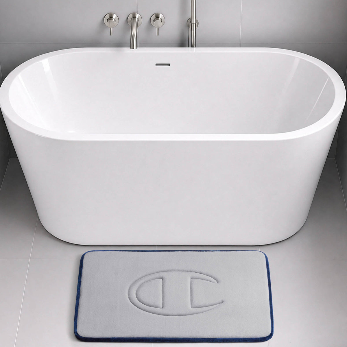 A modern white bathtub with wall-mounted faucets sits in a grey tiled bathroom. In front of the tub is a light grey Champion Quilted Logo Memory Foam Bath Mat, featuring a raised C logo, plush memory foam comfort, and a dark blue border.