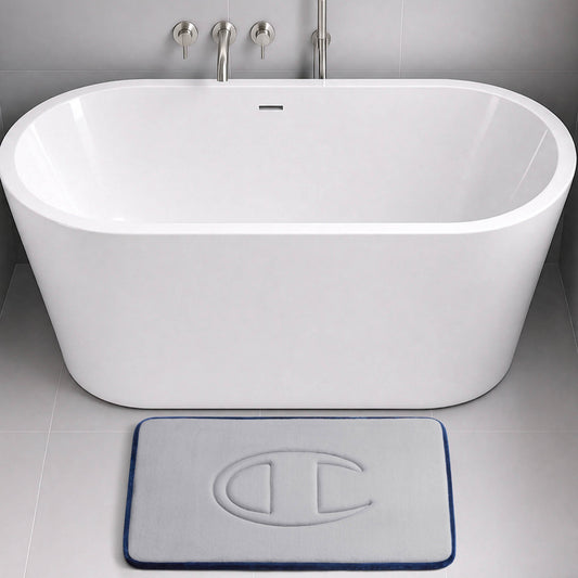 A modern white bathtub with wall-mounted faucets sits in a grey tiled bathroom. In front of the tub is a light grey Champion Quilted Logo Memory Foam Bath Mat, featuring a raised C logo, plush memory foam comfort, and a dark blue border.