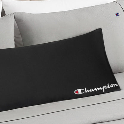 A black Champion Jersey Body Pillow featuring the iconic logo and script rests on a bed dressed in light gray pillows and sheets adorned with small Champion logos, creating a sporty, athletic accent.