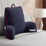 A dark gray Champion Faux Wool Backrest Pillow with red piping rests on a neatly made bed. Made from soft faux wool, the pillow offers supportive arms, a convenient side pocket, and sports a Champion label. A lamp and part of a nightstand can be seen next to the bed.