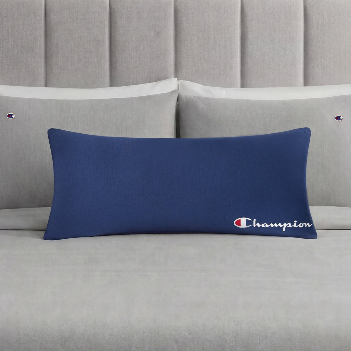 A Champion Jersey Body Pillow in bold blue rests on a neatly made bed with light gray Champion-branded pillows and sheets. A light gray tufted headboard enhances the understated sporty vibe of the room.