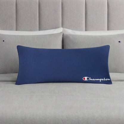 A Champion Jersey Body Pillow in bold blue rests on a neatly made bed with light gray Champion-branded pillows and sheets. A light gray tufted headboard enhances the understated sporty vibe of the room.