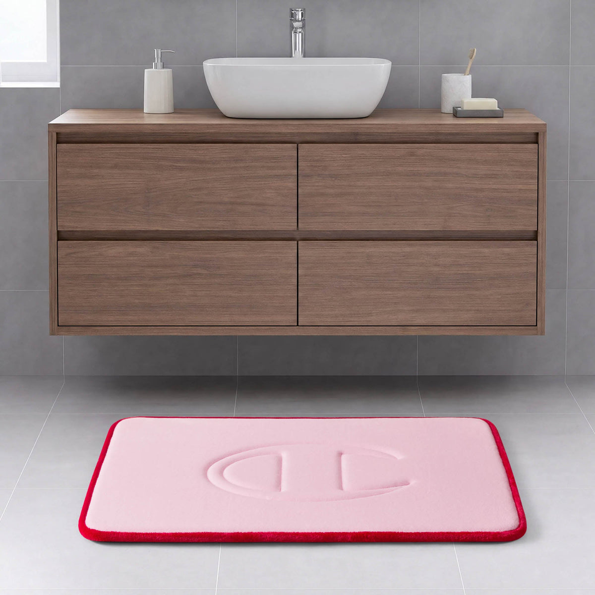 A modern bathroom with a wooden floating vanity, a white rectangular basin, and the Quilted Logo Memory Foam Bath Mat by Champion—featuring a pink surface with a red border and an embossed design—placed on a light gray tiled floor.