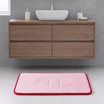 A modern bathroom with a wooden floating vanity, a white rectangular basin, and the Quilted Logo Memory Foam Bath Mat by Champion—featuring a pink surface with a red border and an embossed design—placed on a light gray tiled floor.