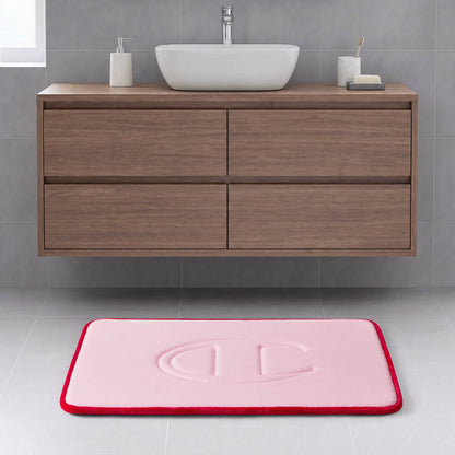 A modern bathroom with a wooden floating vanity, a white rectangular basin, and the Quilted Logo Memory Foam Bath Mat by Champion—featuring a pink surface with a red border and an embossed design—placed on a light gray tiled floor.
