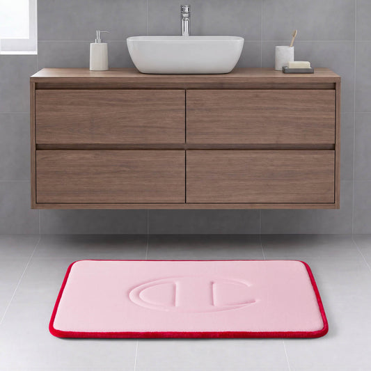 A modern bathroom with a wooden floating vanity, a white rectangular basin, and the Quilted Logo Memory Foam Bath Mat by Champion—featuring a pink surface with a red border and an embossed design—placed on a light gray tiled floor.