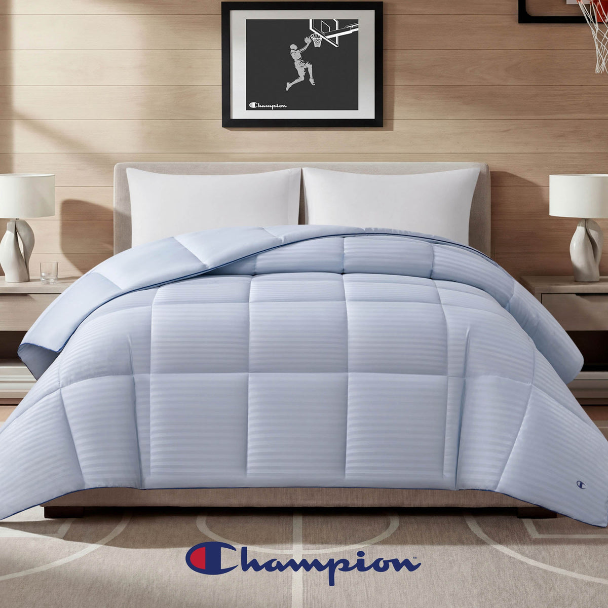 A neatly made bed showcases the Champion Blue Cooling Down Alternative Comforter, complemented by two pillows, nightstands with lamps, and a distinctive Champion logo at the foot of the bed. Basketball-themed framed art hangs on the wall behind, completing this stylish setup featuring Champion bedding.