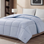 A Blue Cooling Down Alternative Comforter by Champion is displayed on a bed in a modern bedroom with white pillows, a lamp, bookshelf, and framed picture on the wall. The space features neutral decor and soft lighting.
